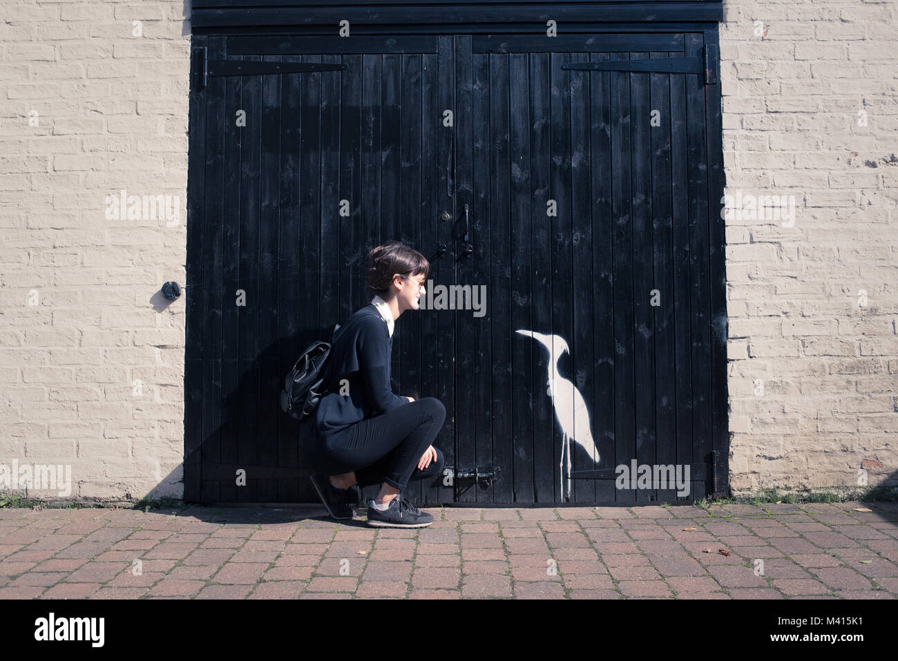 Hipster Frau mit schwarzen und Sonnenbrille auf der Suche nach einem kleinen weißen Vogel Zeichnung (ähnlich wie ein Reiher) auf einem großen schwarzen Holz Garagentor Stockfoto