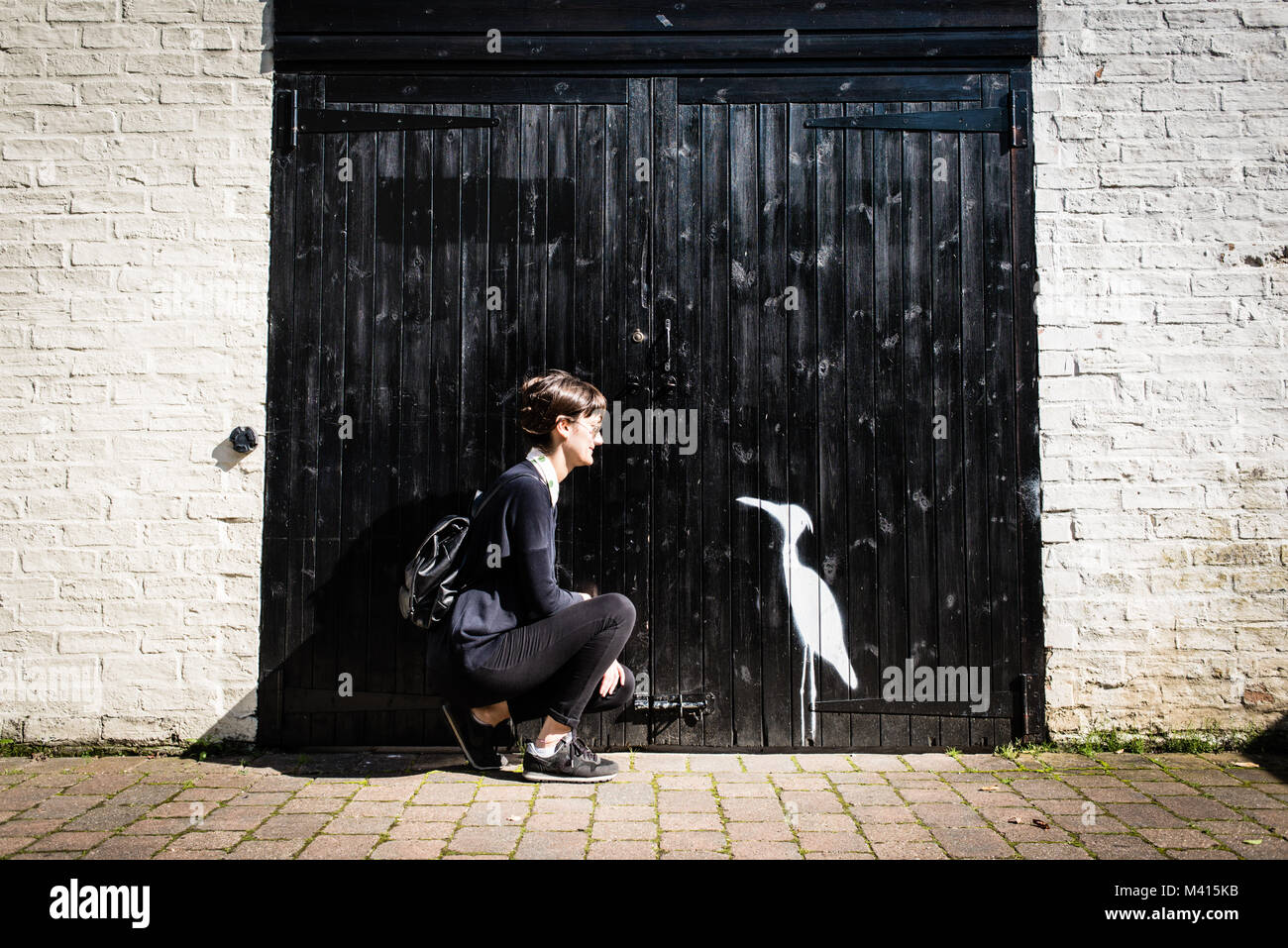 Hipster Frau mit schwarzen und Sonnenbrille auf der Suche nach einem kleinen weißen Vogel Zeichnung (ähnlich wie ein Reiher) auf einem großen schwarzen Holz Garagentor Stockfoto