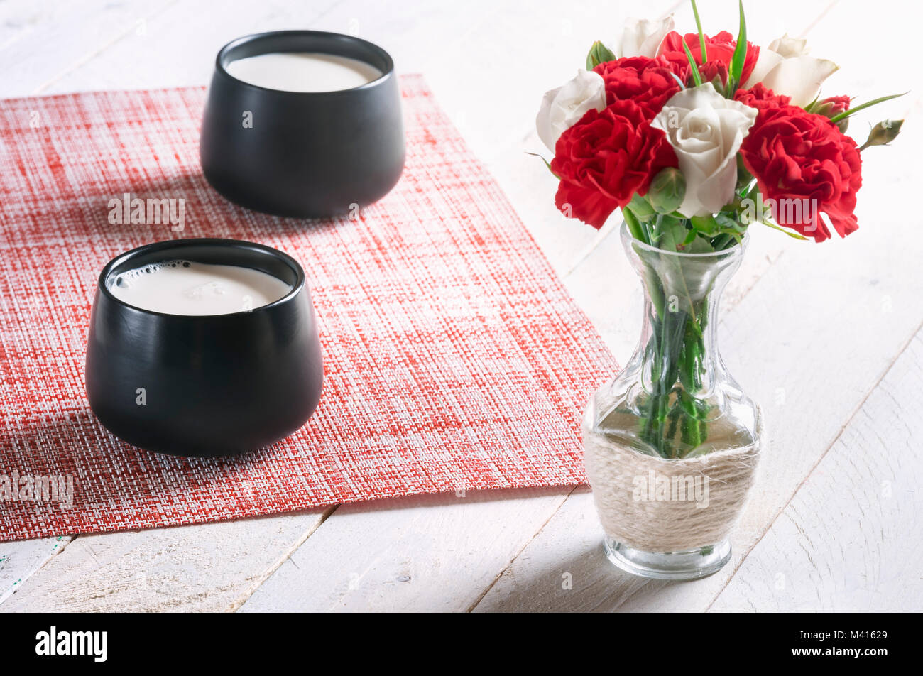 Frühstück Tisch mit zwei schwarze Becher voller Frische Milch und ein Blumenstrauß aus den weißen Rosen und roten Nelken, auf einem weißen Tisch, an einem sonnigen Morgen. Stockfoto