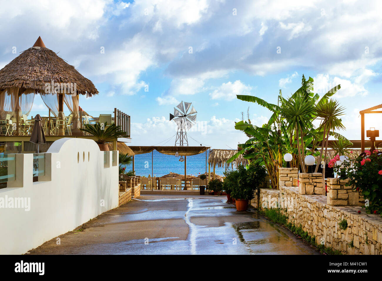 Dekorative Windmühle mit weißen Propeller auf thatched Dach installiert. Tische mit strohgedeckten Sonnenschirme am Strandcafé am Meer. Livadi Strand im Meer, die Bucht Stockfoto