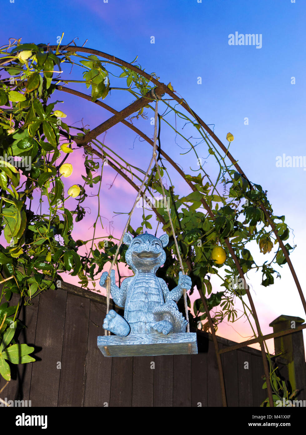 Nett, lächelnd Drachen garten Ornament auf einer Schaukel, vor einen Bogen mit wachsenden Passionsblume / Passiflora, gegen einen schönen Sonnenaufgang Sonnenaufgang. UK. Stockfoto