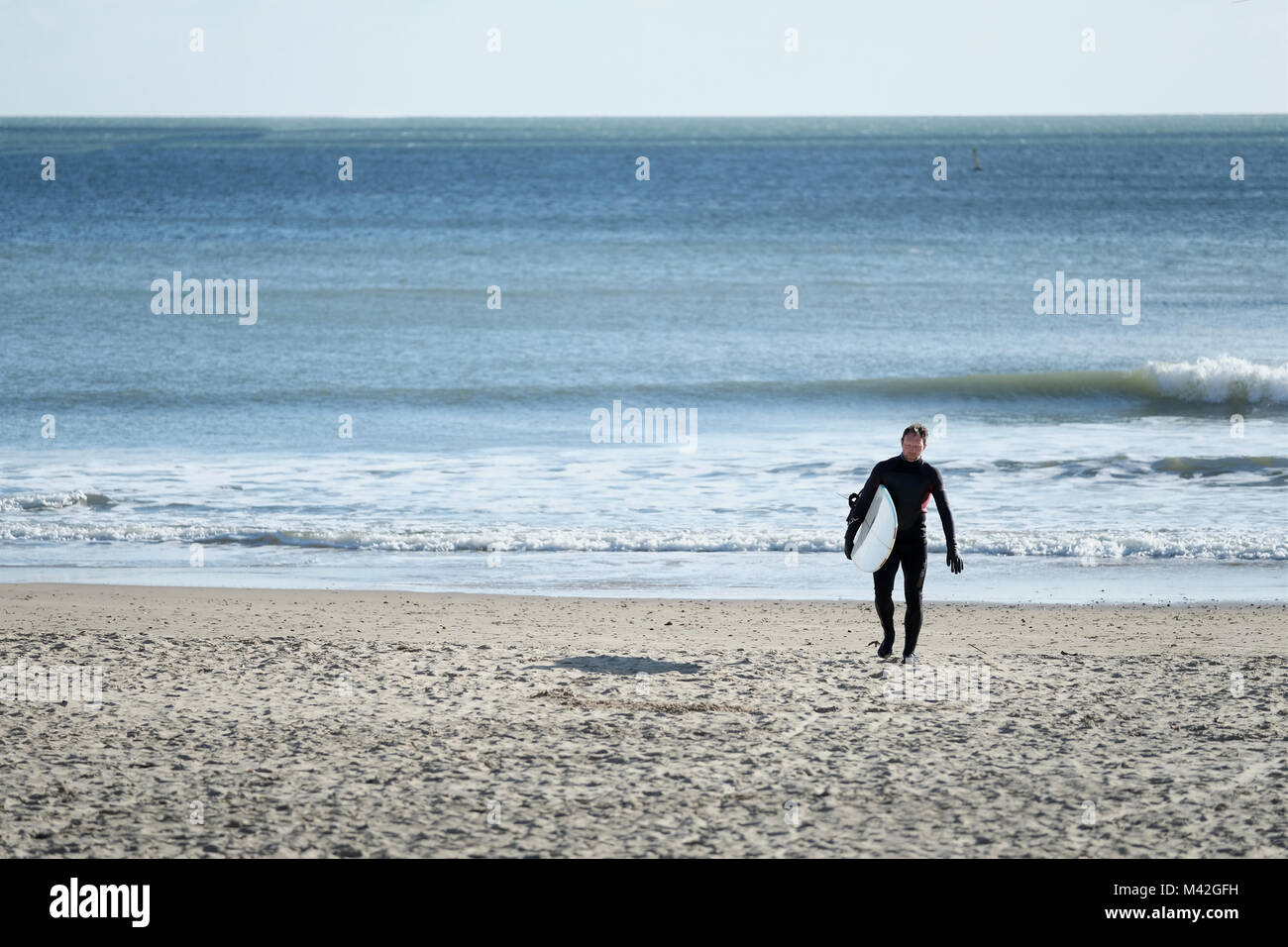 Ein Surfer tragen eine Ganzkörper Anzug, Spaziergänge am Strand, vom Meer, mit seinem Surfbrett auf einem kalten helle Winter Tag in Bournemouth, Großbritannien Stockfoto