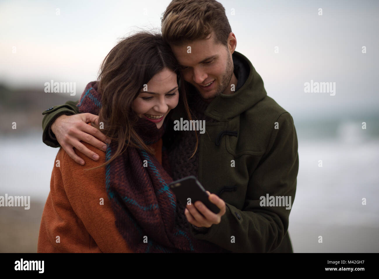 Junge erwachsene Paare an Smartphone suchen am Strand Stockfoto