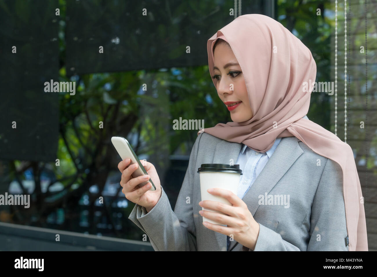 Ziemlich elegante weibliche muslimische Büroangestellter mit Handy Kontrolle der Unternehmens-E-Mail, wenn Sie stand vor der Fenster im Freien entspannen. Stockfoto