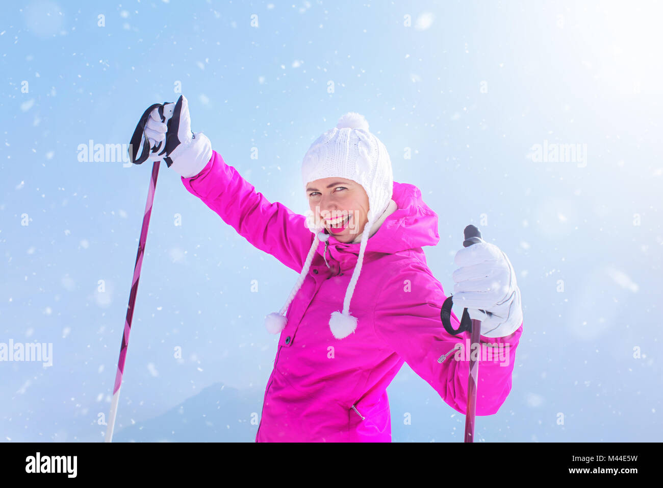 Glückliche junge Frau in rosa Winterjacke, Skistöcke, Handschuhe, lächelnd mit Sonne und Schnee im Hintergrund. Stockfoto
