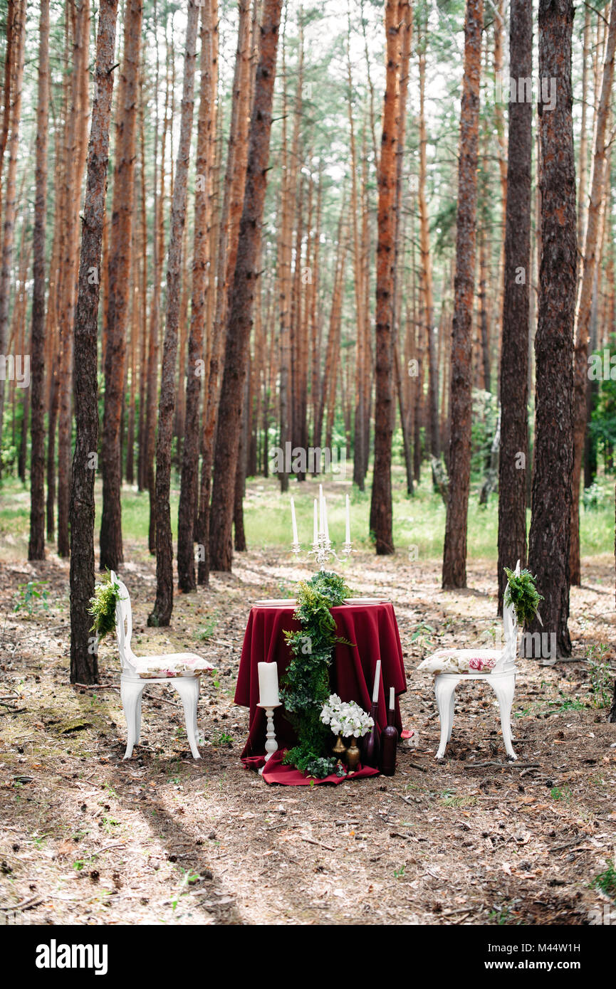 Jäten Dekor. Gedeckter Tisch für zwei serviert mit zwei weißen geschmiedet Stühle im Pinienwald. Dekoration mit Marsala Stoff, Kerzen, Kerzenhalter, Wein Stockfoto