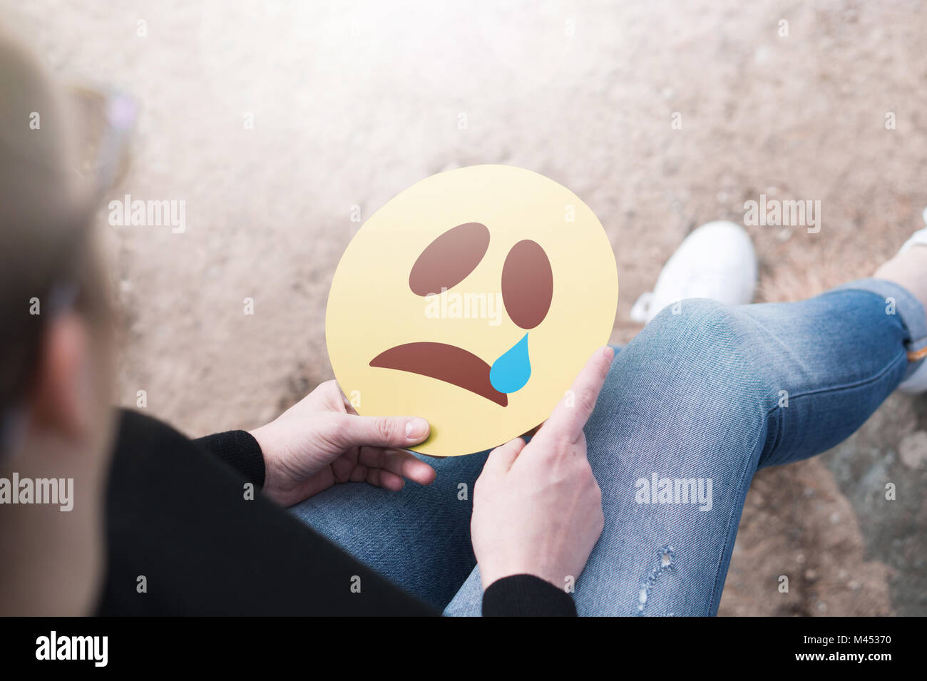 Weinend Papier emoticon in der Hand. Deprimiert Frau mit gedruckten traurige smiley Gesicht und sitzt auf einem Felsen. Moderne Kommunikations- und Smiley. Stockfoto