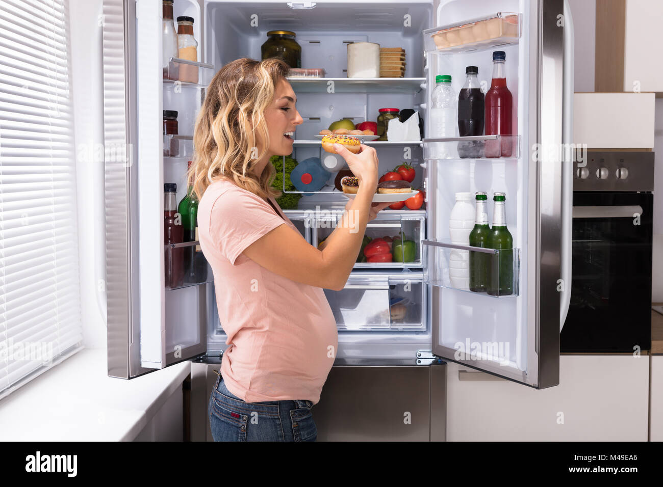 Junge schwangere Frau glücklich essen Donut Vor dem offenen Kühlschrank und in der Küche Stockfoto