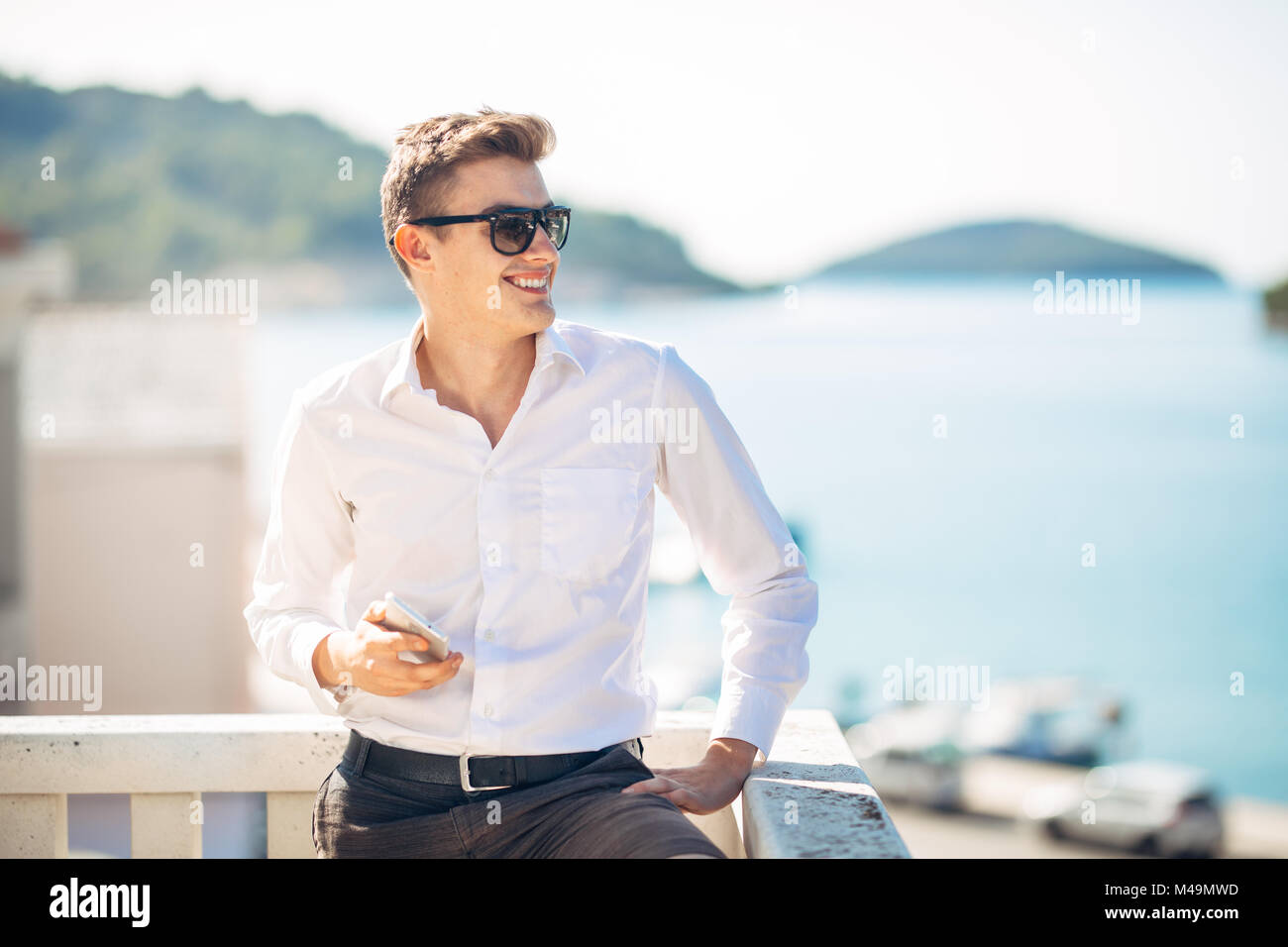 Junger stattlicher Mann mit Aufenthalt im Luxus Resort Hotel mit Panoramablick auf das Meer. lächelnd Geschäftsmann mit seinem Smartphone Optionen zu einem verdienten t Stockfoto