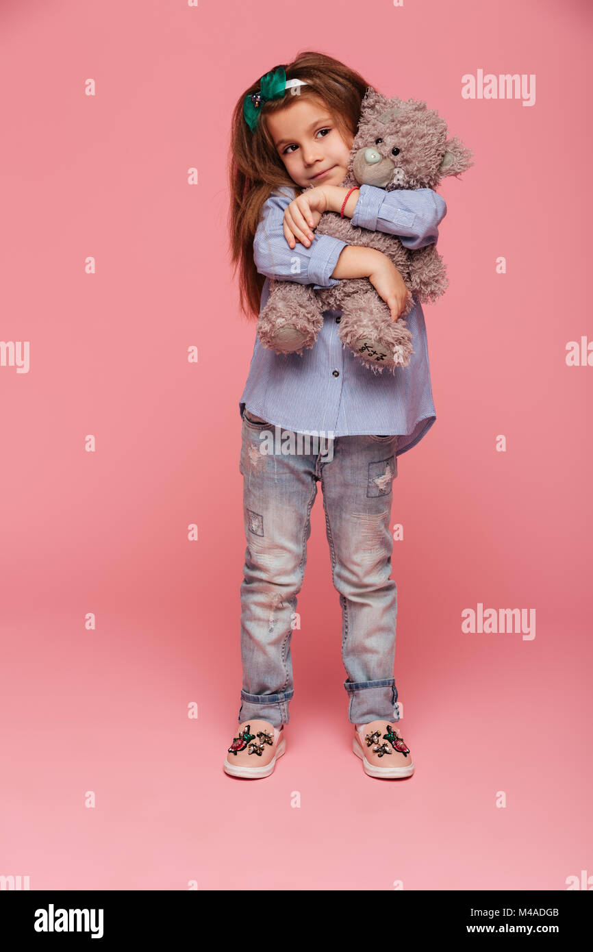 In voller Länge Foto von adorable Kind Mädchen mit langen auburn Haar und legere Kleidung, ihren schönen Teddybär über rosa Hintergrund isoliert umarmen Stockfoto