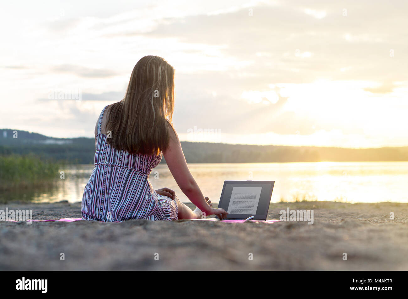 Junge attraktive Frau mit Laptop am Strand bei Sonnenuntergang. Aktiver Lebensstil. Schüler Hausaufgaben machen oder Unternehmer spät. Stockfoto