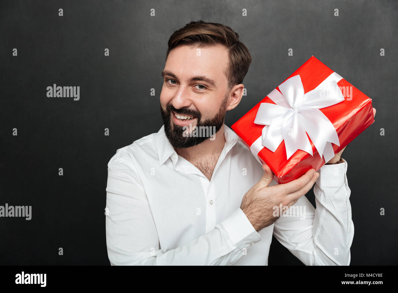 Portrait von neugierigen Menschen schütteln Red Box Geschenk verpackt und versuchen, zu erkennen, was im Inneren über dunkle graue Wand Stockfoto