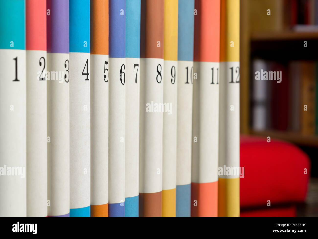 12 nummerierte Bücher in einer Reihe mit einem Bücherregal und ein rotes Sofa im Hintergrund Stockfoto
