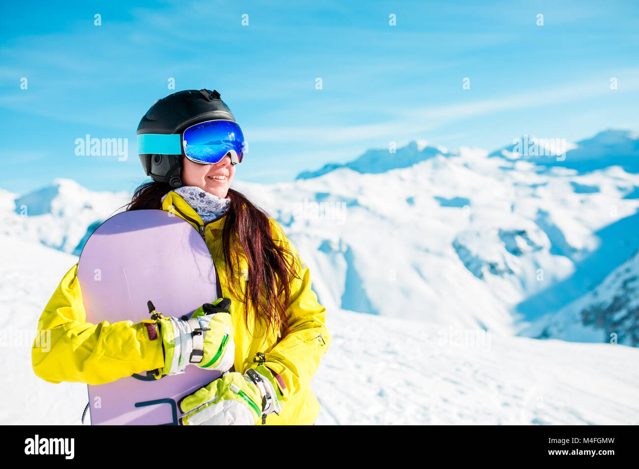 Bild von Smiling brunette das Tragen von Helm und Maske mit Snowboard auf den Hintergrund der schneebedeckten Hügeln bei Tag Stockfoto