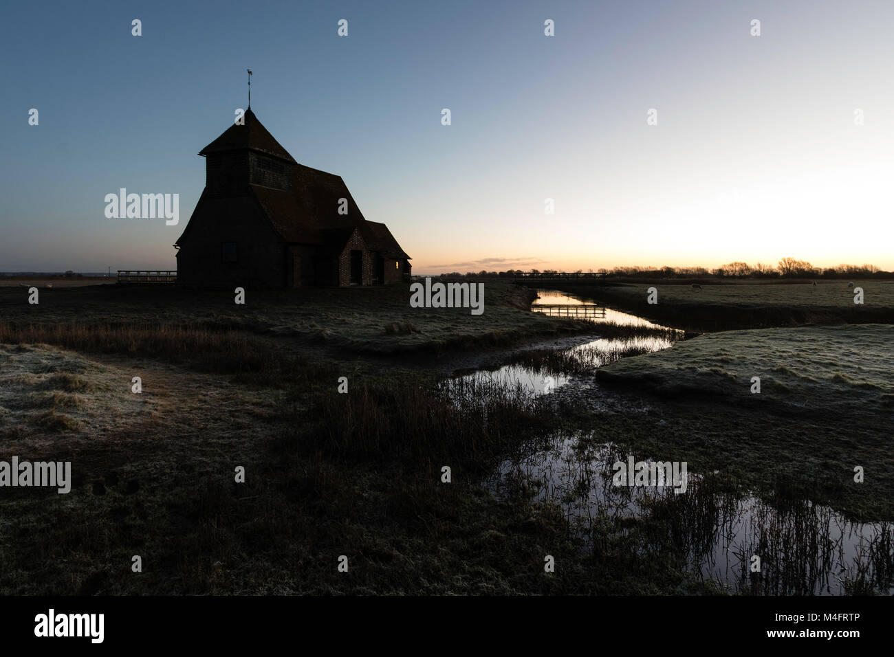 Der hl. Thomas Becket Kirche in die Sümpfe. Winter Dawn Twilight, Frost auf den Boden, halb gefrorenen Graben mit Schilf, Kirche am Horizont. Romney Marsh, Großbritannien. Stockfoto