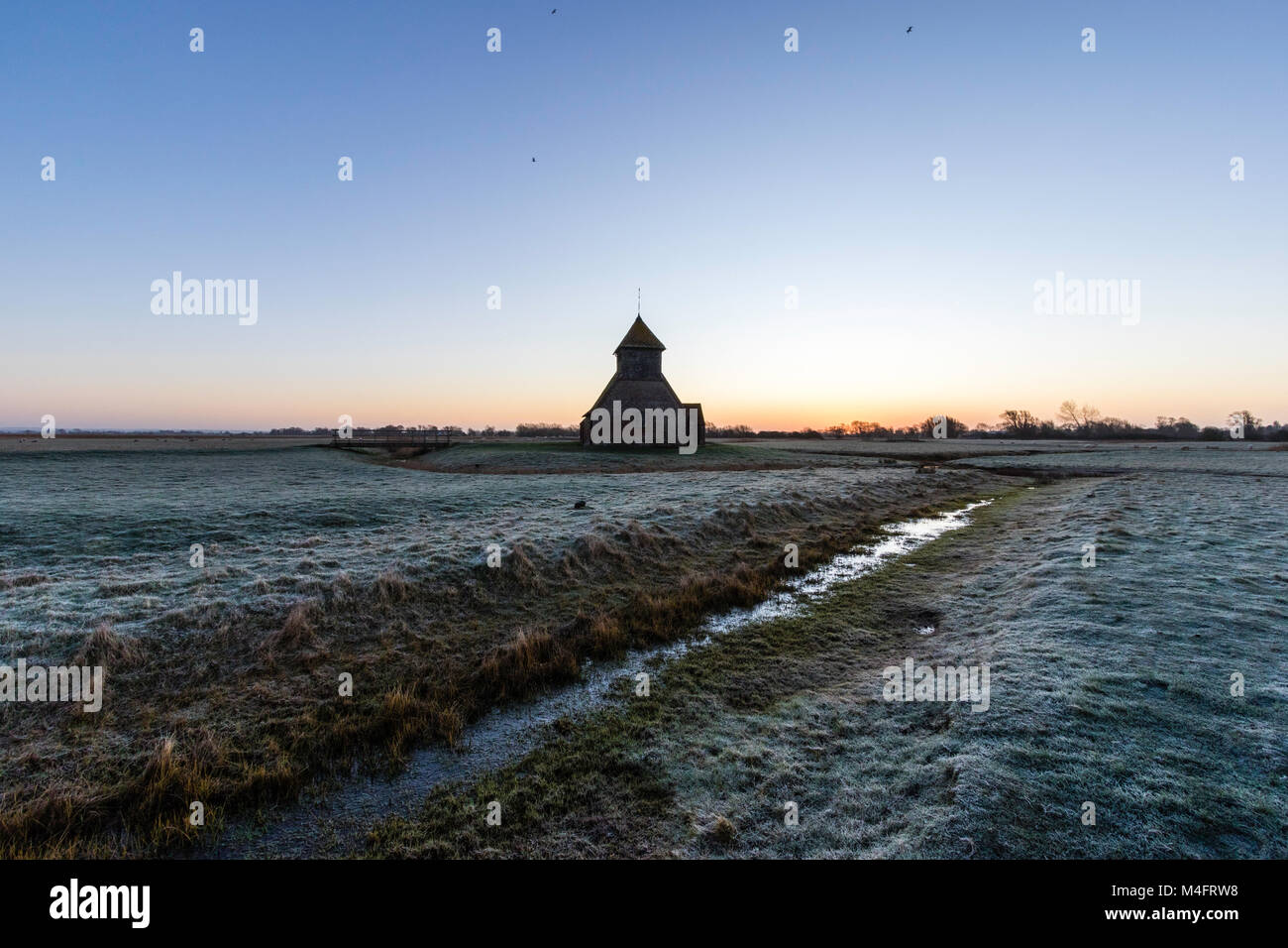 Der hl. Thomas Becket Kirche in die Sümpfe. Winter Dawn Twilight, Frost auf den Boden, halb gefrorenen Graben mit Schilf, Kirche am Horizont. Romney Marsh, Großbritannien. Stockfoto