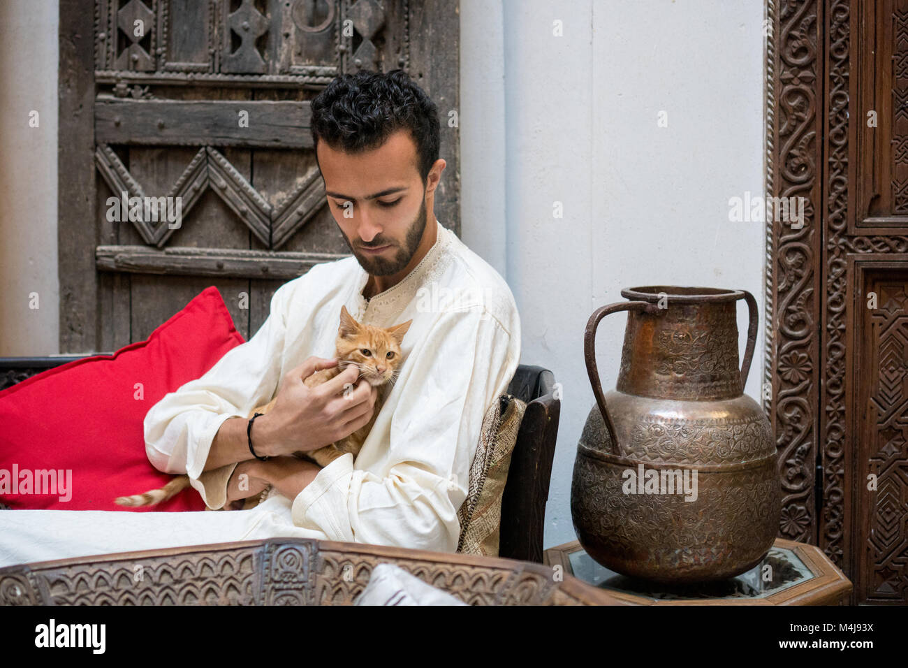 Junge muslimische Mann in traditioneller Kleidung, die eine Gelbe Katze vor einem geschmückten Wand Stockfoto