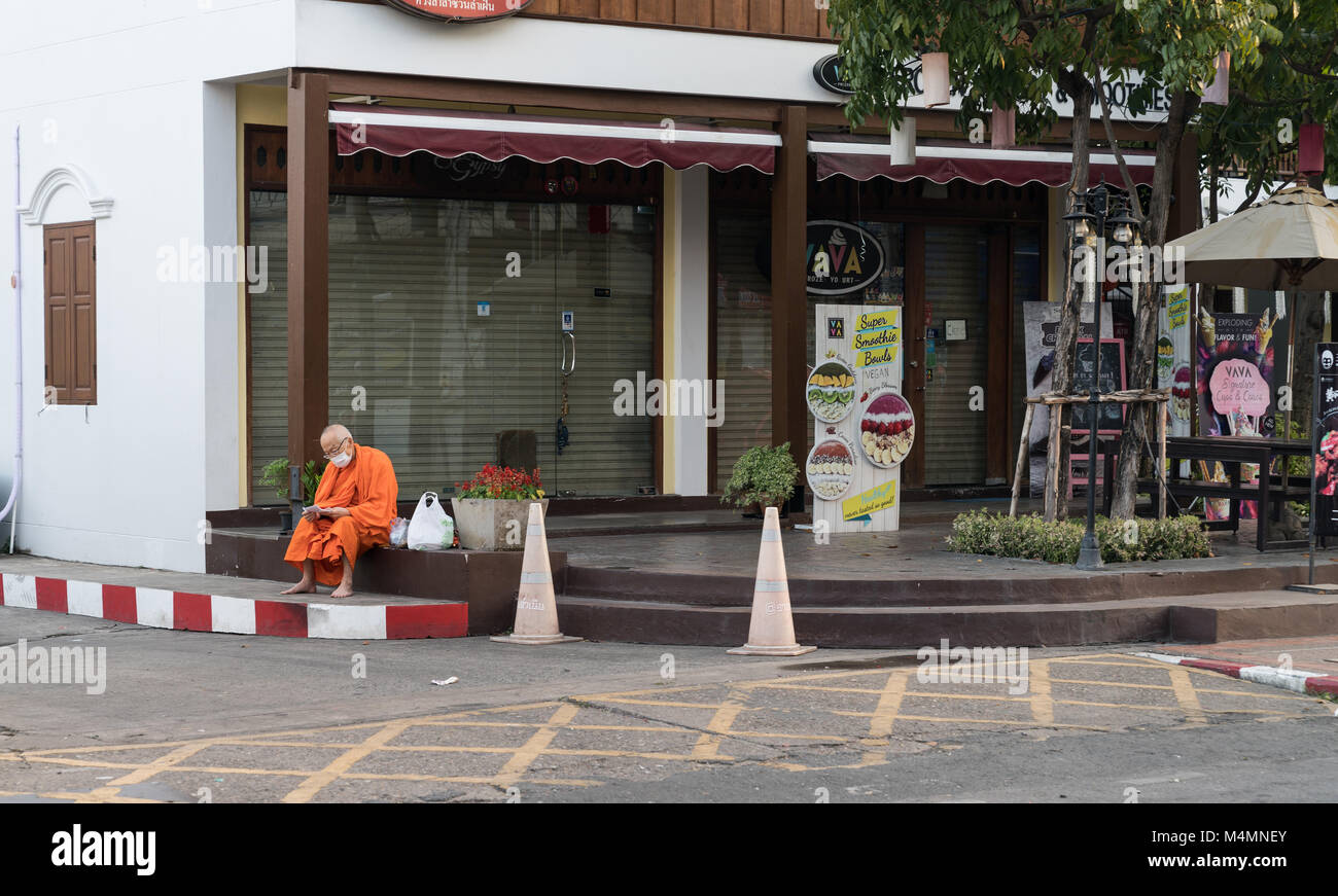 CHIANG MAI, THAILAND - 29. Januar 2018; Mönch in Safran Roben tragen von Op-Gesichtsmaske mit plstic Einkaufstüten sitzt auf Ecke Pflaster außen Stockfoto