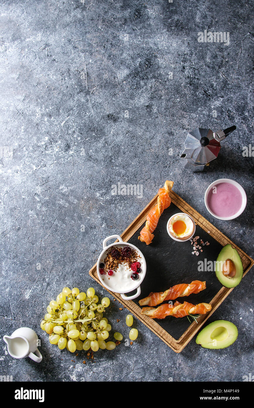 Frühstück mit geräuchertem Lachs auf knusprige Grissini, Joghurt, Smoothie, Avocado, Müsli, Milch, Beeren und grüne Trauben über blaue Textur Hintergrund gesetzt. Stockfoto