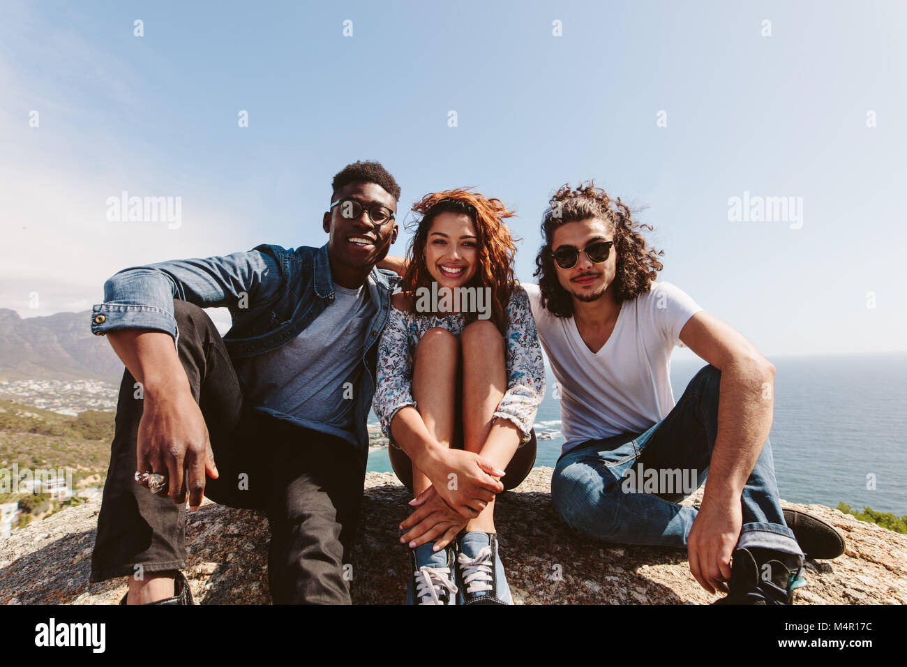 Eine Gruppe von Freunden auf einem Berg. Lächelnde junge Leute zusammen sitzen draußen auf der Spitze einer Klippe. Stockfoto