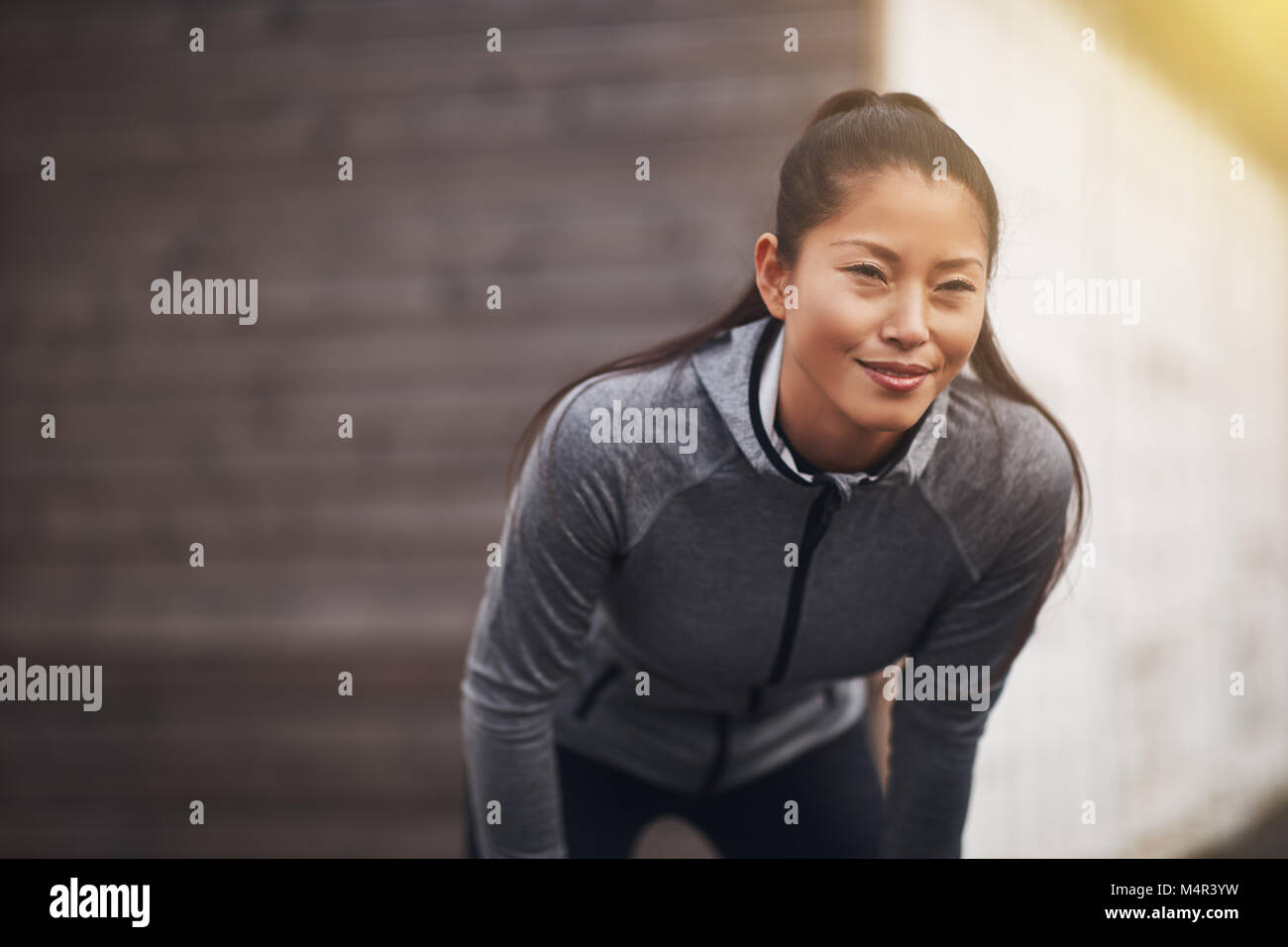 Lächelnde junge asiatische Frau stehend außerhalb übung Kleidung tragen, mit den Händen auf den Knien die Vorbereitung für einen Lauf zu gehen Stockfoto