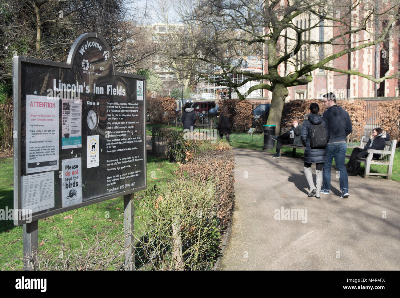 Menschen sitzen und Spaziergang auf einem Februar Tag in Lincoln's Inn Fields, London, England, neben einem Empfang und Information Board Stockfoto