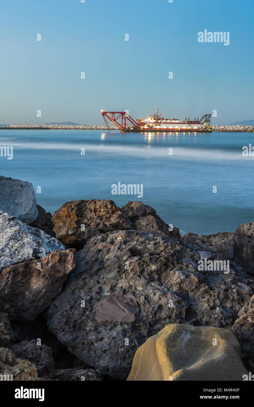 Bagger Boot, geführt von der Morgen leuchtet im Hafen arbeiten mit Felsbrocken im Vordergrund. Stockfoto