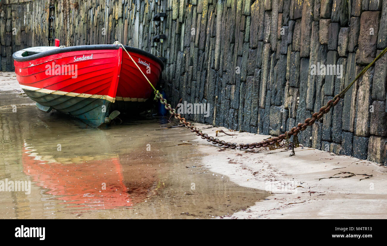 Nahaufnahme eines angeketteten kleinen roten Ruderbootes bei Ebbe im malerischen Hafen von Portsoy, Aberdeenshire, Schottland, Großbritannien, mit Wasserspiegelungen Stockfoto