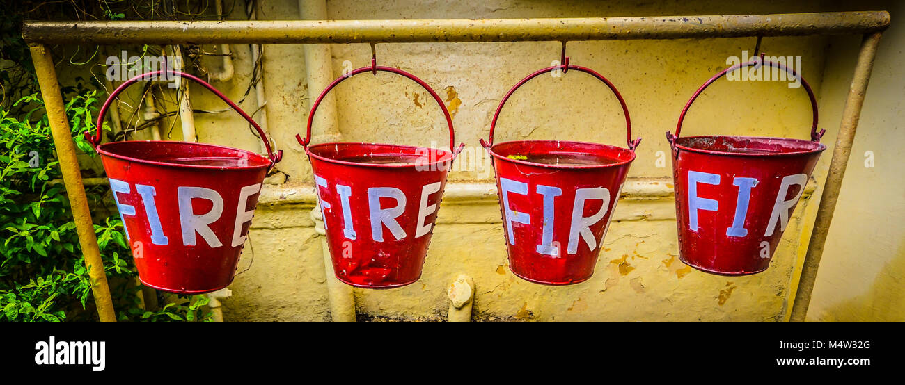Eine Reihe von vier roten Eimer mit der Bezeichnung "Feuer" in weißen Buchstaben für das Sammeln von Wasser ein Feuer gegen gelbe Wand mit grünen Sträuchern und Reben zu begießen. Stockfoto