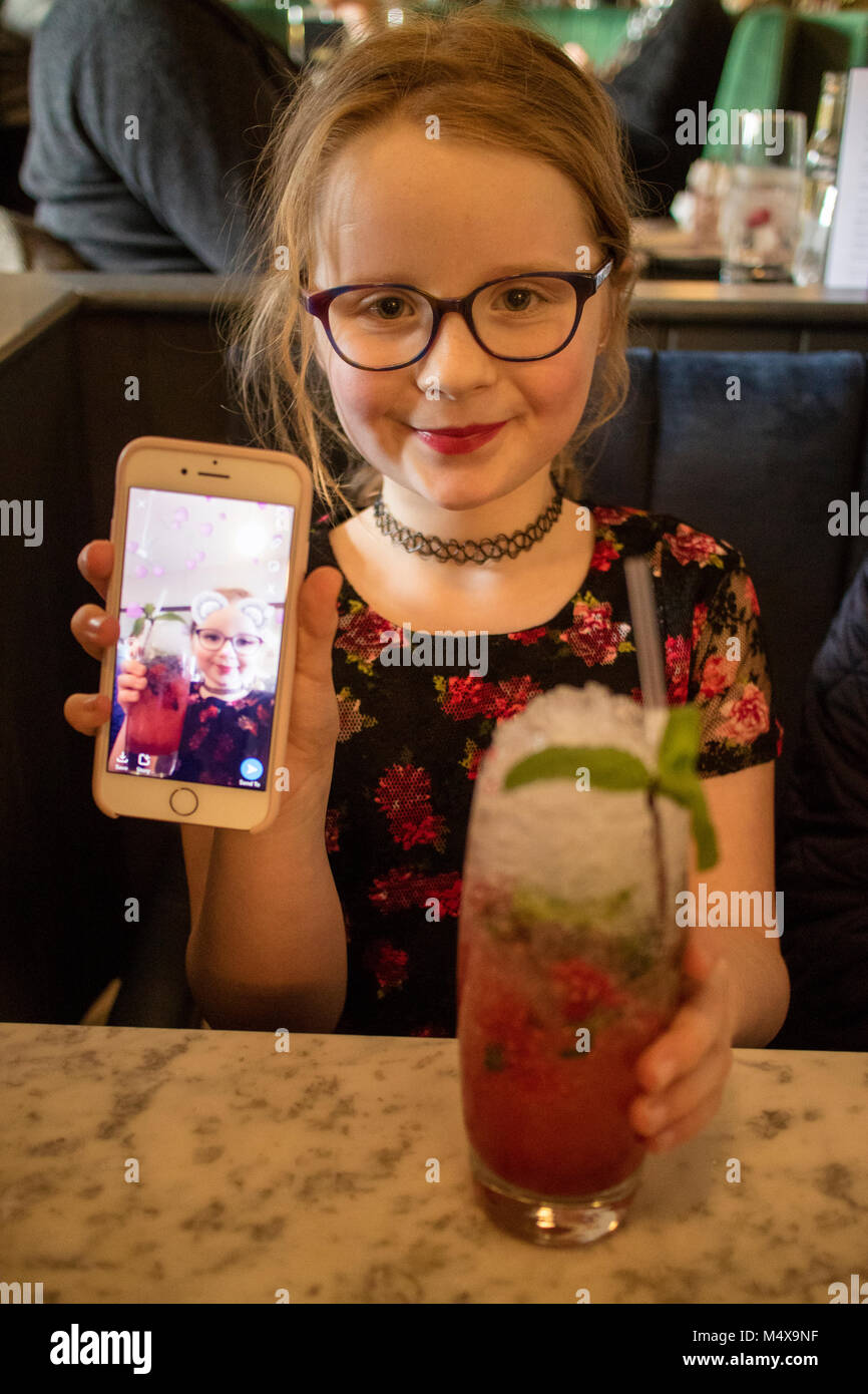 8-Jähriges Mädchen zeigen snapchat auf Ihrem iPhone Smartphone während des Sitzens in einer Bar trinken einen alkoholfreien Cocktail Stockfoto