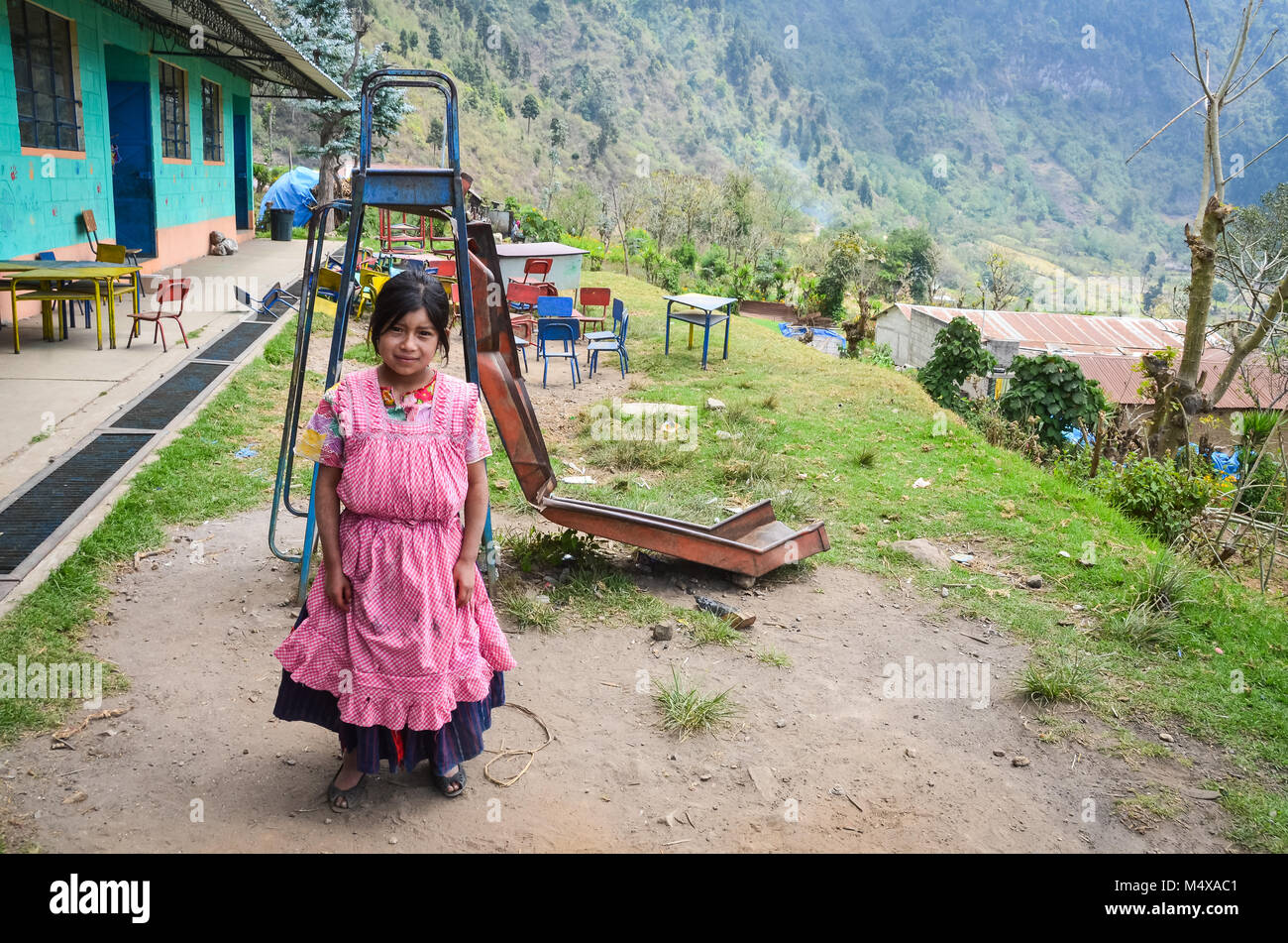 Eine guatemaltekische Mädchen in Ethik Kleid steht vor einem zerbrochenen Rutsche auf dem Spielplatz in einer armen ländlichen Schule. Stockfoto
