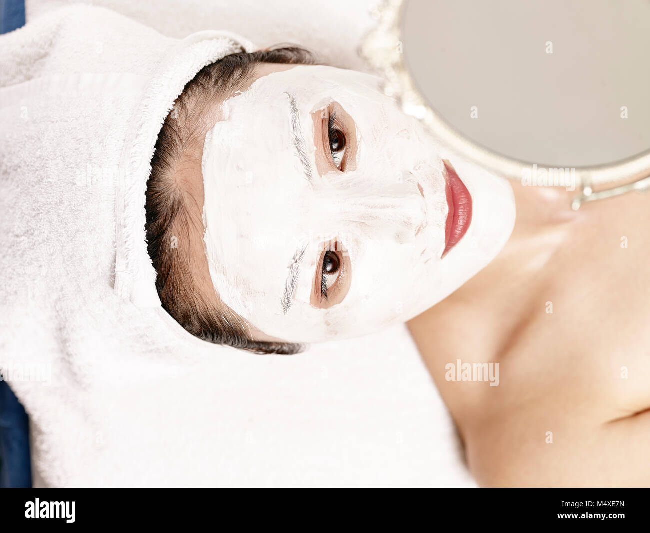Schöne junge asiatische Frau mit Gesichtsmaske lag auf dem Rücken auf dem Bett Blick in den Spiegel. Stockfoto