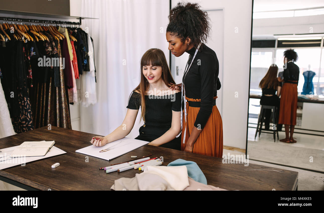 Fashion Designer Skizzieren eines Design sitzen an Ihrem Tisch, während eine andere Frau auf aussieht. Mode Unternehmer diskutieren ein Kleid Design in ihrer Art Stockfoto