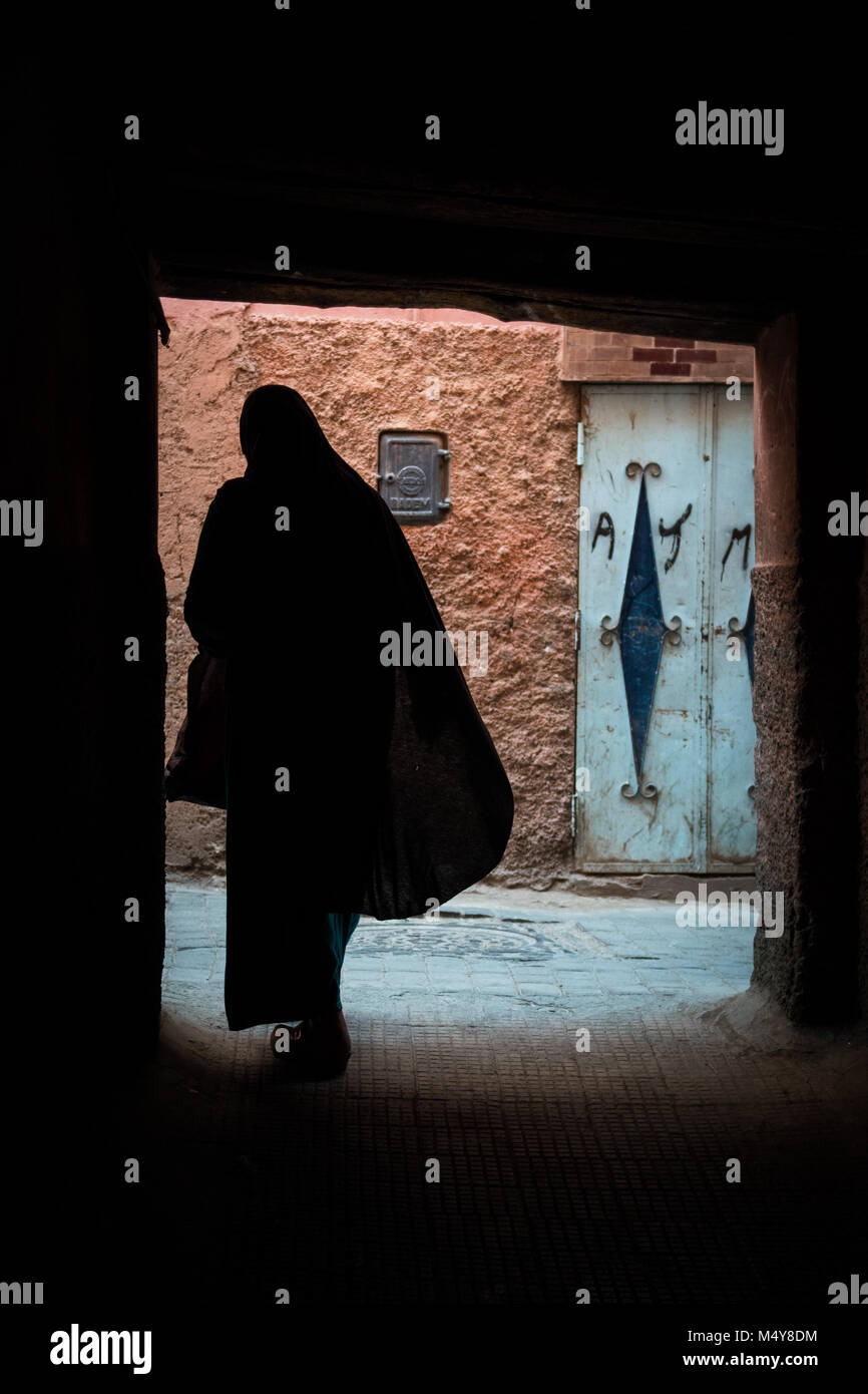 Eine dunkle Silhouette einer marokkanische Frau in traditioneller Kleidung gegen einen hellen Steinmauer. Die Mauer ist Pfirsichfarben mit einem grünen blauen Tür. Stockfoto