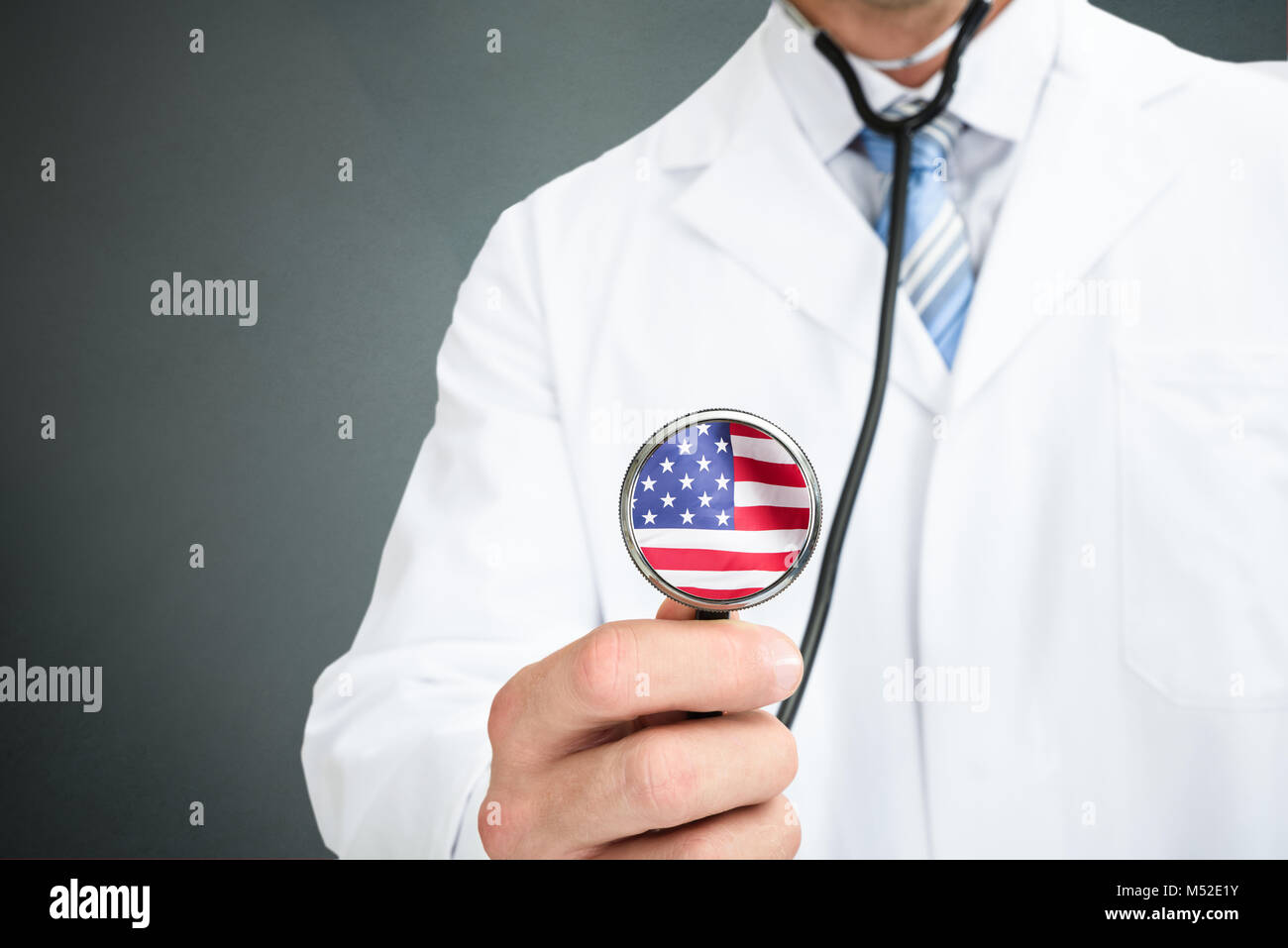 Mittelteil der männlichen Arzt Holding Stethoskop mit amerikanischer Flagge auf Chestpiece gegen grauer Hintergrund Stockfoto