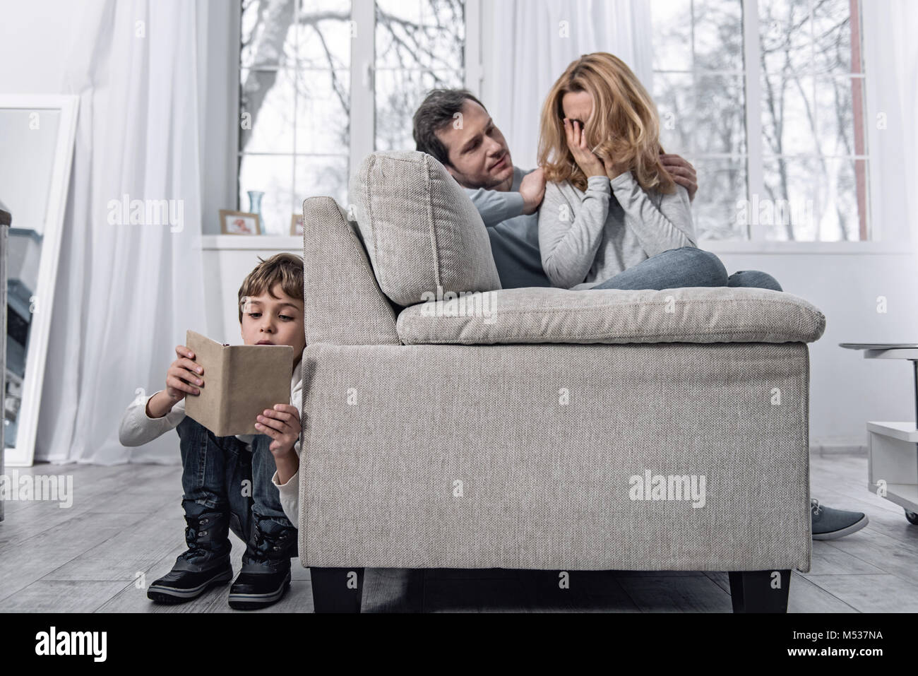 Junge Frau weint und ihr Sohn ein Buch lesen Hinter dem Sofa Stockfoto