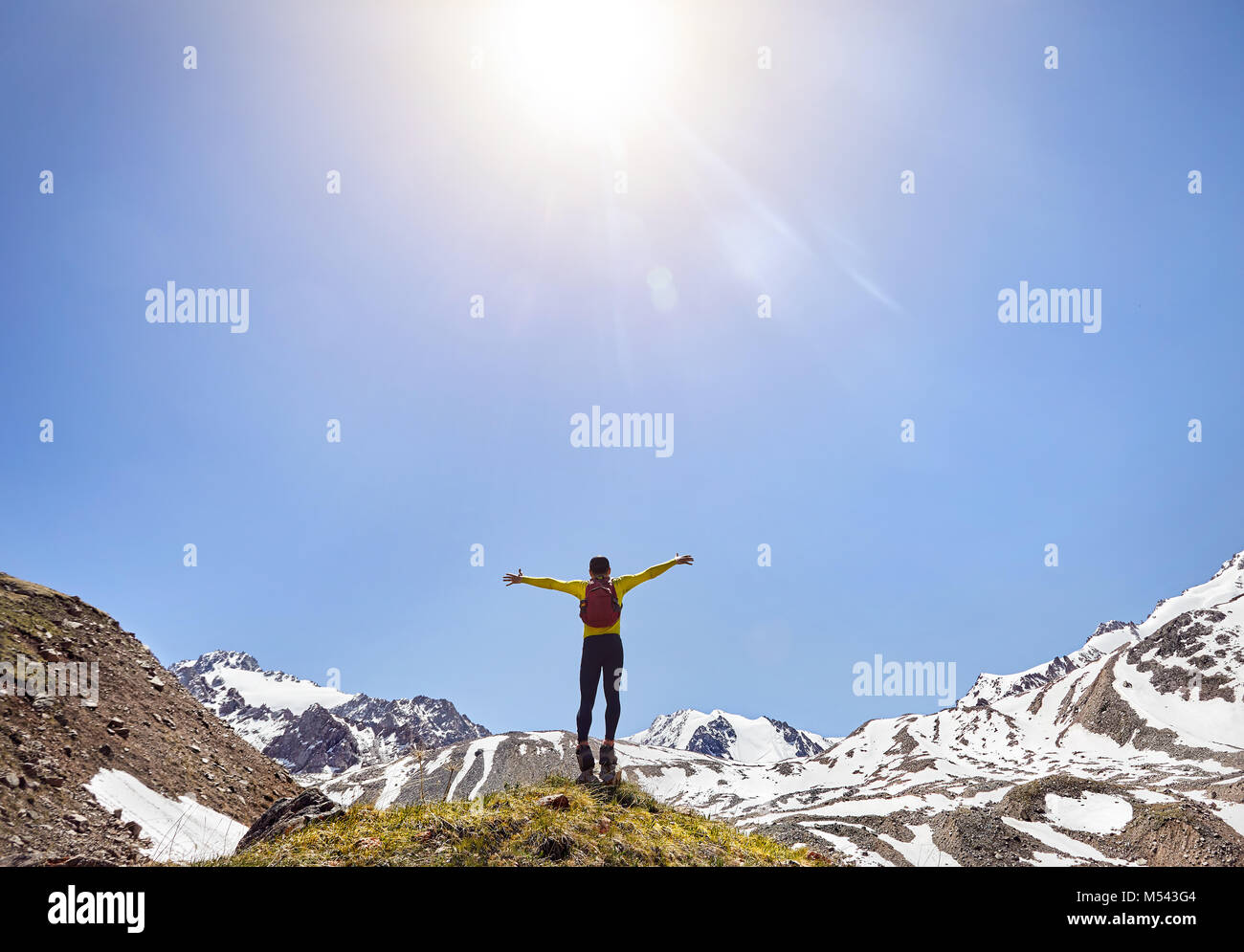 Wanderer in gelb Shirt mit Rucksack auf den Felsen mit steigenden Hände genießen den Blick auf schneebedeckte Berge bei Sunny day Sky Stockfoto