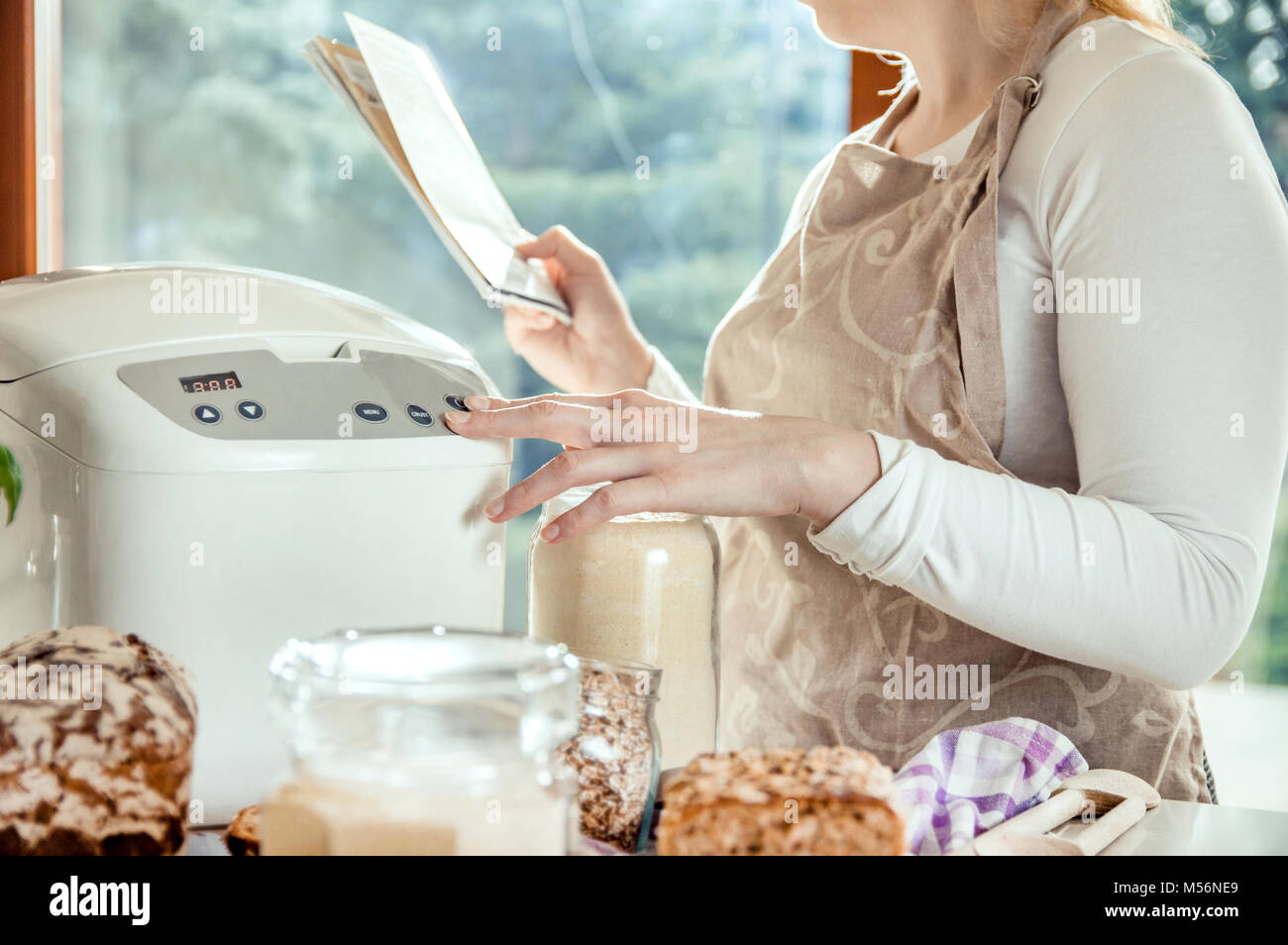 Frau, die in einer modernen Küche liest die Anweisungen für eine Maschine, die das Brot backt. Foto im Licht der natürlichen Sonne und Studio Lampen genommen. Es enthält Stockfoto