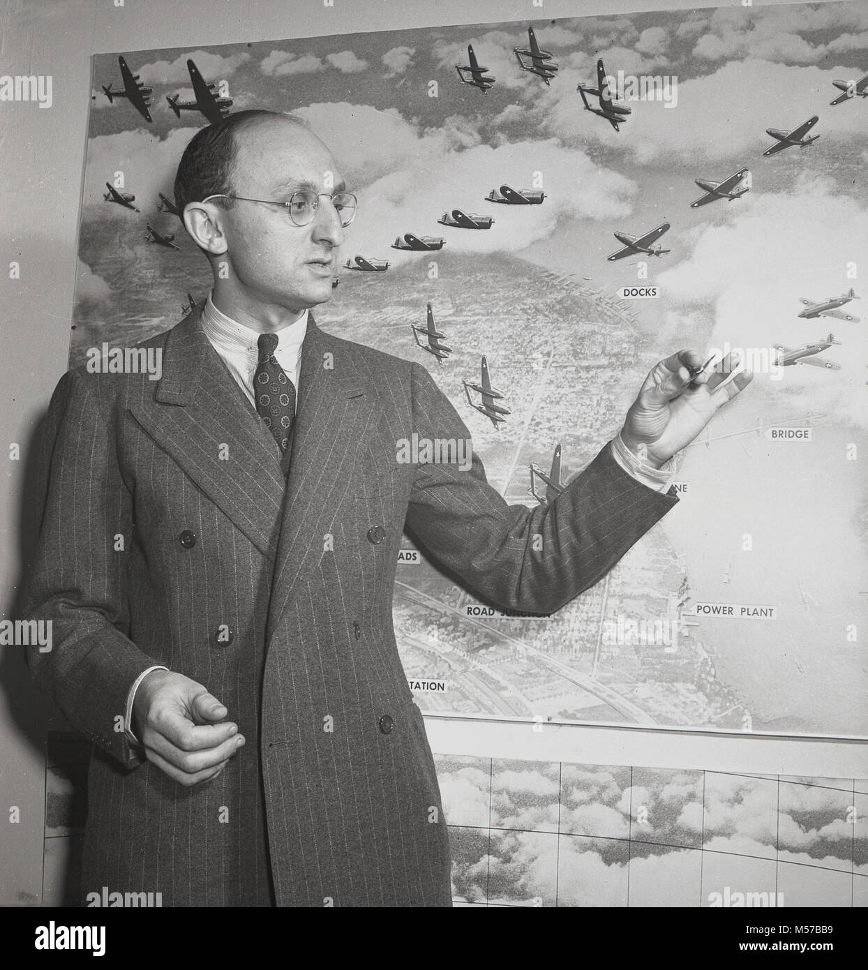1942, USA, historische Bild, ein Mann mit einem Doppel-suit breasted stehen und Erklärung "Air Combat" durch eine große Zeichnung an der Wand von Flugzeugen und strategischen Ziel, Schauplätze, wie zum Beispiel Docks und Kraftwerke bombardiert werden. Stockfoto