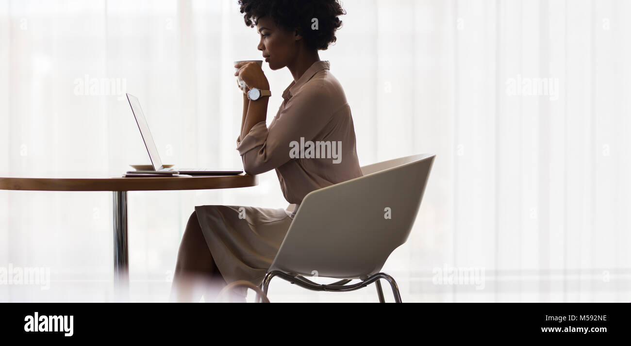 Afrikanische Geschäftsfrau, trinken Kaffee und Laptop auf dem Tisch. Seitenansicht der weiblichen Executive mit Laptop und Kaffee im Büro. Stockfoto