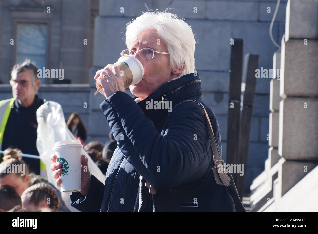 Eine ältere Frau, trinken ein heißes Getränk von einem verwerfbaren Schale in warme Kleidung mit Menschen im Hintergrund gekleidet, Trafalger Square, London Stockfoto