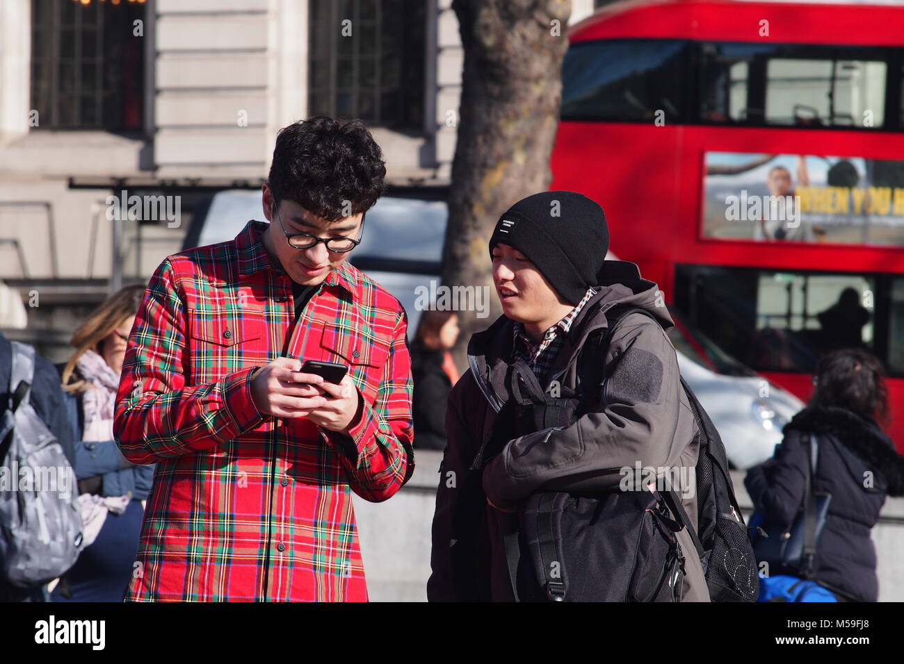 Zwei junge Männer auf der Suche nach einem Smartphone im Winter Sonnenschein, Trafalgar Square, London mit einem Doppeldeckerbus im Hintergrund Stockfoto