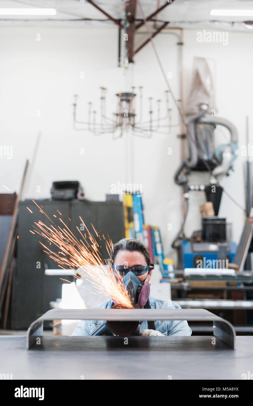 Frau das Tragen von Schutzbrille und Staubmaske stehend in der Metallwerkstatt, mit der Schleifmaschine, Funken fliegen. Stockfoto