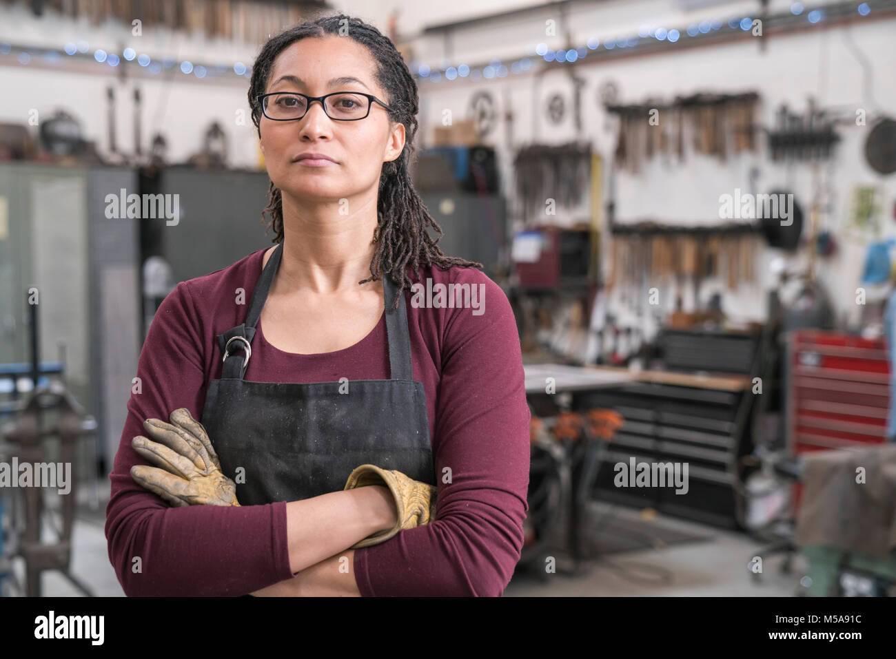 Frau mit braunem Haar Brille und Schürze in der Metallwerkstatt, die Arme verschränkt, an der Kamera schaut. Stockfoto