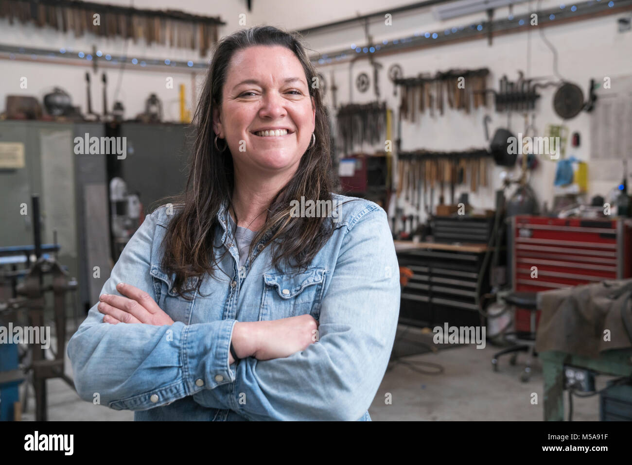 Frau mit braunem Haar zu tragen Jeanshemd in der Metallwerkstatt, lächeln in die Kamera. Stockfoto