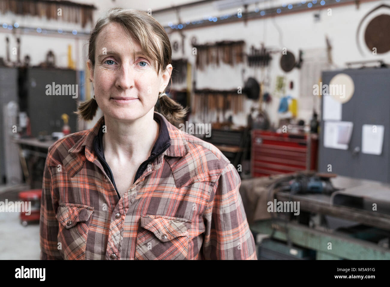 Blonde Frau mit kariertem Hemd in der Metallwerkstatt, an der Kamera schaut. Stockfoto