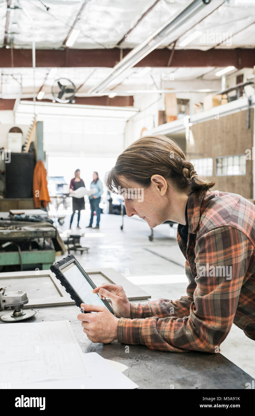 Seitenansicht der blonden Frau mit kariertem Hemd an Workbench in der Metallwerkstatt, Holding digital Tablet. Stockfoto