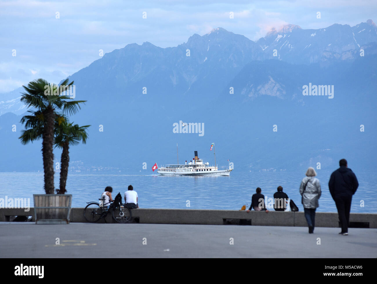 Die Menschen am Ufer am Genfer See in Lausanne mit Bootsfahrt und die Berge im Hintergrund. Stockfoto