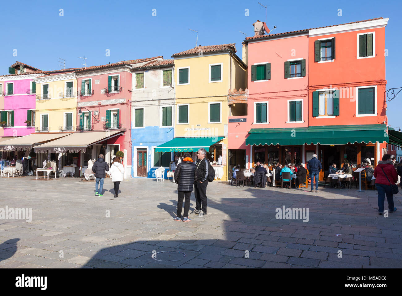 Insel Burano, Venedig, Venetien, Italien. Bunte Geschäfte und Restaurants mit Menschen speisen in der Via Baldassare Galuppi im Winter Stockfoto