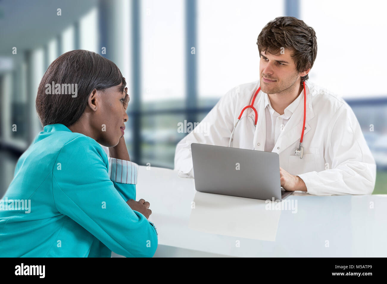 Männlicher Arzt in einem Gespräch mit Ihrem african american, Patienten, während Sitzen am Tisch in der Nähe der Fenster im Krankenhaus. Arzt ist bereit, Patienten zu helfen. Sitzen auf den großen hospitawindows Büro Stockfoto